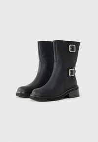 Botines de cuero negro con una textura suave, punta redonda, dos acentos de hebilla metálica y un taco grueso para mayor estabilidad.