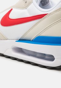 Tenisová obuv s bílým mesh svrškem, béžovými překryvy, červeným Swooshem a modrým akcentem na podrážce. Obsahuje viditelnou vzduchovou jednotku a texturovanou černou podrážku.