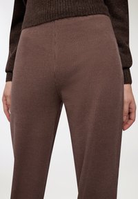 Zbliżenie na osobę w brązowych, teksturowanych spodniach i ciemniejszym brązowym swetrze, z rękami swobodnie opuszczonymi przy bokach.