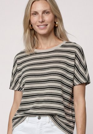 Femme blonde portant des boucles d'oreilles créoles dorées et un haut beige à manches courtes avec des rayures noires horizontales, rentrée dans un pantalon blanc.
