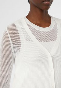 Weißer gestrickter Cardigan mit einem Netzdesign, V-Ausschnitt und Knopfschluss. Weiche Textur mit einem geschichteten Look darunter.