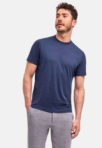 Man met krullend haar en baard, gekleed in een marineblauw t-shirt en lichtgrijze broek, staand met zijn handen in zijn zakken tegen een effen achtergrond.