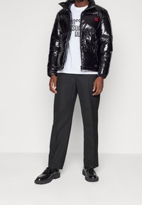 Czarna błyszcząca kurtka puchowa z wysokim kołnierzem, zestawiona z białą koszulką z grafiką. Szare spodnie i czarne buty na platformie dopełniają look.