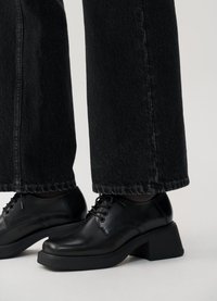 Sapatos oxford pretos em couro com bico arredondado e salto bloco grosso, combinados com jeans escuros de perna larga com detalhe de costura na bainha.
