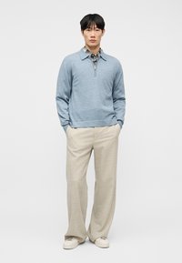 Νεαρός άνδρας που στέκεται με τα χέρια στις τσέπες, φορώντας ανοιχτό μπλε πουλόβερ με φερμουάρ πάνω από καρό πουκάμισο, μπεζ φαρδιά παντελόνια και λευκά αθλητικά παπούτσια.