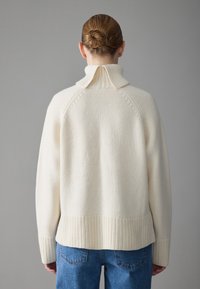 Pull en tricot crème avec un col haut, des finitions côtelées et des poignets. Présente une coupe décontractée et un tissu texturé.