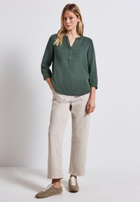 Blusa verde scuro texturizzata con scollo a V e maniche a tre quarti, abbinata a pantaloni beige a gamba dritta e sneakers marrone chiaro.