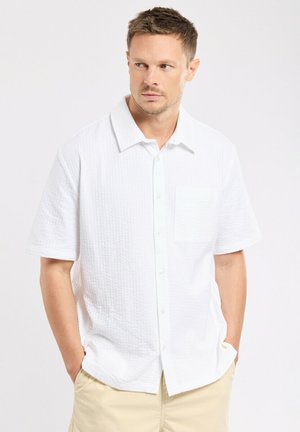 Homme portant une chemise blanche texturée à manches courtes avec poche, pantalon beige, mains dans les poches, regardant légèrement vers la gauche sur un fond uni.