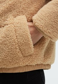 Beige fleece jacket with a textured surface, ribbed cuffs, and elastic waistband. Features side pockets for added functionality.