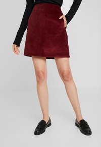 Gonna in corda bordeaux con una forma svasata, dotata di tasche laterali e una texture liscia. Abbinata a mocassini neri per contrasto.