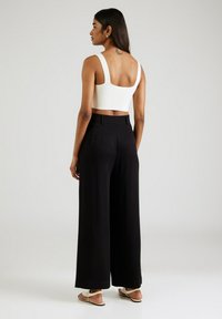 Femme de dos portant un top court blanc, un pantalon large noir, des sandales blanches, avec un tatouage dans le haut du dos.