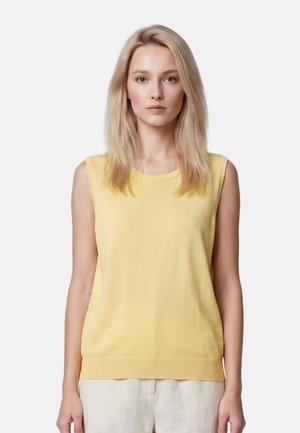 Giovane donna con capelli biondi lisci indossa un top giallo senza maniche e pantaloni chiari, in piedi contro uno sfondo chiaro e uniforme.