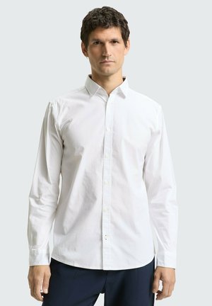 Man draagt een effen wit overhemd met lange mouwen en donkere broek, staand tegen een lichtgrijze achtergrond.