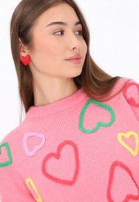 Różowy sweter z dzianiny z wielokolorowymi haftowanymi sercami w kolorze czerwonym, zielonym, żółtym i fioletowym. Uzupełniony czerwonymi kolczykami w kształcie serca.