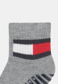 Calzino in cotone grigio con bordo a costine, decorato con un motivo geometrico in blu, bianco e rosso, e dettagli in gomma nera antiscivolo sulla suola.