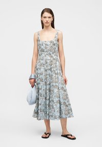 Γυναίκα που φοράει αμάνικο, μπλε φόρεμα μήκους μίντι με λουλούδια, μαύρα σαγιονάρες και κρατά μια ανοιχτό μπλε τσάντα με φαρδιά λαβή, τυλιγμένη στον καρπό της.