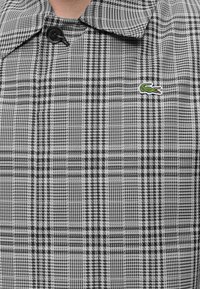 Veste à carreaux noir et blanc avec col pointu, bouton noir et petit logo crocodile vert sur la poitrine.