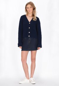 Cardigan lavorato a maglia blu navy con scollatura a V e cinque bottoni dorati, abbinato a una gonna di jeans scura e sneaker bianche. Tessuto testurizzato, vestibilità rilassata.