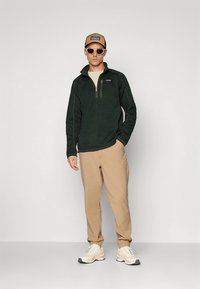 Mörkgrön fleece-tröja med halv dragkedja, tanfärgade byxor, beige keps och vita sneakers; solglasögon kompletterar den avslappnade outfiten.