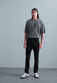 Junger Mann mit langen dunklen Haaren, der ein übergroßes graues T-Shirt, schwarze Jeans und weiße Sneaker trägt, steht vor einer schlichten grauen Wand.