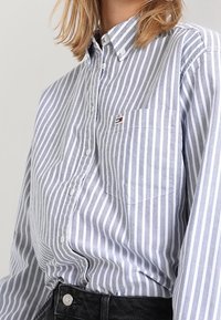 Camicia a righe blu e bianche con bottoni, dotata di taschino sul petto, realizzata in tessuto morbido. Presenta un piccolo patch con logo sulla tasca.