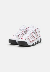 Vitaidrottsskor i vitt med röda accenter och perforerade grå paneler. Tjock svart gummisula och framträdande "AIR"-logotyp på sidorna.