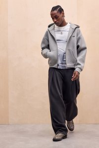 Sweat à capuche gris zippé avec une grande poche avant, superposé à un t-shirt blanc à motif, associé à un pantalon noir ample plissé et des chaussures à motifs.