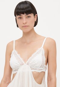 Biely bralet s čipkovým lemovaním, tenkými ramienkami, výstrihom do tvaru V a mäkkou, priesvitnou látkou na spodnej časti.