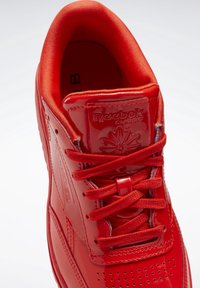 Czerwone, błyszczące sneakerzy Reebok Classic z pasującymi sznurowadłami, szczegółowym przeszyciem i markowanym językiem, sfotografowane z góry na białym tle.