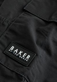Melna auduma ar teksturētu virsmu, ar taisnstūrveida melnu etiķeti, uz kuras baltā krāsā ir uzraksts "BAKER by TED BAKER".