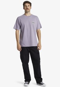 Camiseta de algodón de color púrpura claro con un diseño gráfico en el pecho, combinada con pantalones cargo negros y zapatillas de lona negras.