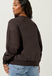 Maglione marrone scuro con vestibilità rilassata, orlo a costine e maniche lunghe, abbinato a jeans azzurri chiari. Tessuto liscio con un design minimale.