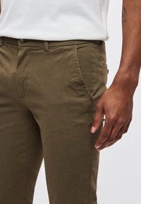 Homme portant un pantalon vert olive et un débardeur blanc côtelé, une main détendue le long de la cuisse.