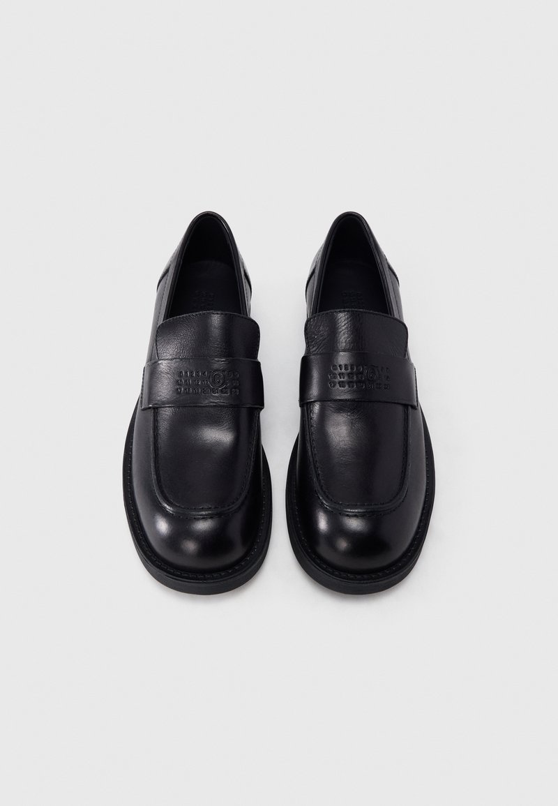 Par af sorte læder slip-on loafers med runde tæer og præget logo på remmen, vist på en ensartet lys baggrund.