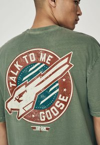 Olivovozelené tričko s kruhovou grafikou, na které je text "Talk to Me Goose" a stylizovaný design letadla, doplněné hvězdami a pruhy.