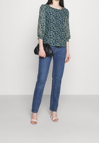 Hedvábná blůza s květinovým vzorem a průhlednými rukávy, v modro-zelených odstínech, kombinována s světle modrými džíny a černou kabelkou, doplněná o bílé sandály na podpatku.