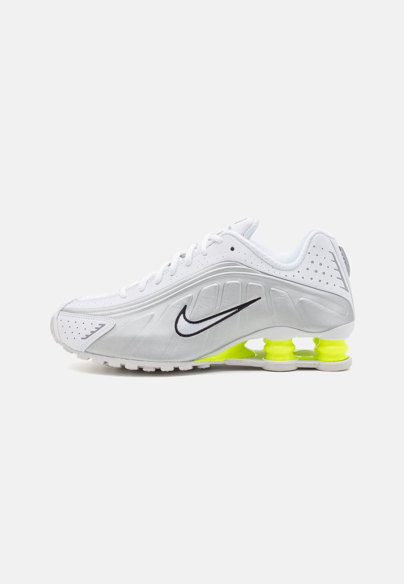 Balti Nike sportbačiai su tekstūruotu viršumi, juodu logotipu ir neoninės geltonos spalvos kulno paminkštinimu. Yra perforacijos, užtikrinančios oro pralaidumą, ir išlenkta forma.