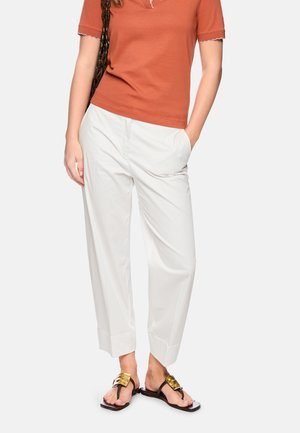 Woman wearing rust orange short-sleeve top, white high-waist cropped pants, black sandals with gold buckles, and black shoulder bag.