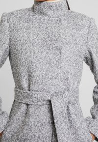 Frau in einem hellgrauen, strukturierten Wollmantel mit hohem Kragen und einem gebundenen Gürtel in der Taille, mit den Händen in den Manteltaschen.