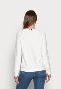 Pull blanc en maille avec un col rond et des poignets côtelés. Présente un petit détail rayé rouge, blanc et marine au dos.