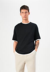 Black short-sleeved t-shirt with white neckline and sleeve trim, made from soft fabric. Model wears beige pants, hands in pockets.