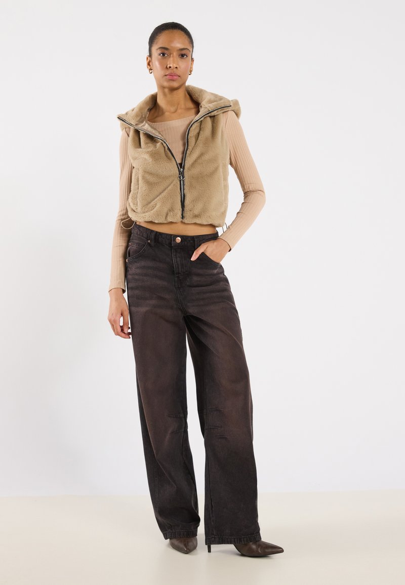 Beige nepleren cropped vest met een capuchon en een zilveren rits, gecombineerd met brede zwarte denim broek en puntige bruine hakken.