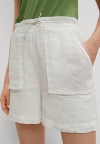 Weißer Leinen-Shorts mit einem Kordelzugbund, Seitentaschen und einer lockeren Passform. Die Textur sieht leicht zerknittert aus, was die Weichheit des Stoffes betont.
