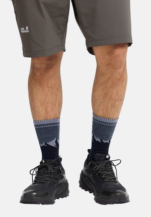 Beine in dunkelgrauer Jack Wolfskin-Shorts, dunkelblauen Socken mit Bergmuster und schwarzen Wanderschuhen mit gemusterten Schnürsenkeln.