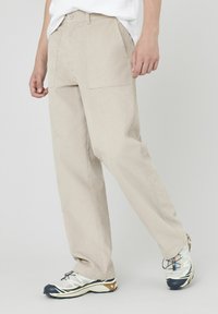 Pantalons beige texturés à coupe décontractée, dotés de deux poches avant et d'une fermeture à bouton unique, associés à des chaussures légères.