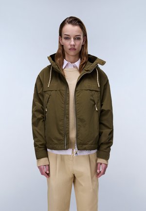 Młoda kobieta o prostych włosach, ubrana w oliwkowozieloną kurtkę z kapturem na beżowym swetrze i jasnych spodniach, zwrócona przodem o neutralnym wyrazie twarzy.