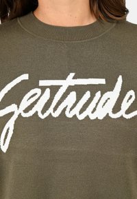 Pull olive en tricot à col côtelé, orné d'un grand texte en tricot blanc "Getstude" en police cursive. Texture lisse, design décontracté.