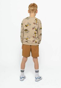 Felpa beige con motivi giocosi, abbinata a pantaloni corti marroni. Presenta polsini e orlo a coste, indossata con calze bianche e scarpe con dettagli blu.