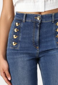 Μπλε τζιν παντελόνια ψηλόμεσα με χρυσά κουμπιά στη αριστερή πλευρά, ελαφρώς ξεθωριασμένα με λεπτομέρειες αντίθεσης στη ραφή.