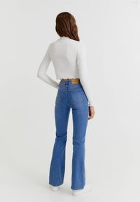 Top blanco corto de manga larga con dobladillo ondulado, combinado con jeans acampanados de tiro alto en azul, que presentan un parche de cuero en la parte trasera de la cintura.